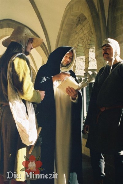 Szene in einem hochmittelalterlichen Kreuzgang: ein Mönch zitiert aus einem Buch. Zwei Handwerker lauschen andächtig.