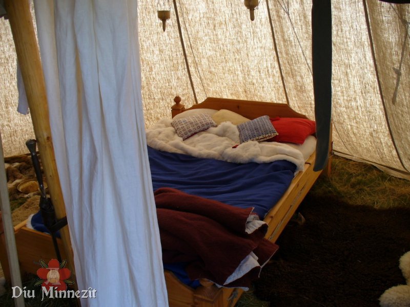 Blick auf ein rekonstruiertes Holzbett in einer hypothetischen Feldlagersituation. Im Hintergrund hängende Tranlampen